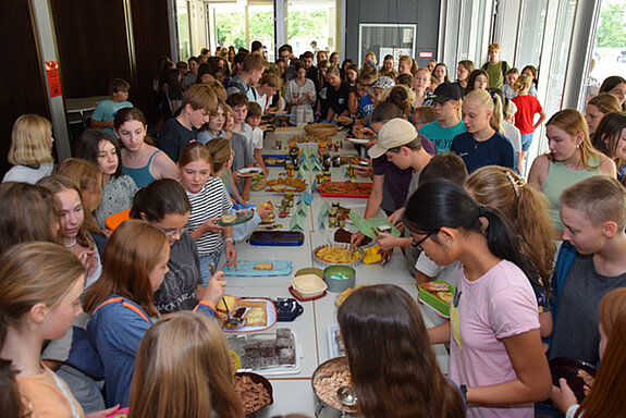 Ein Raum mit vielen Schülerinnen und Schüler, die sich um Tische scharen, auf denen Essen zu sehen ist.