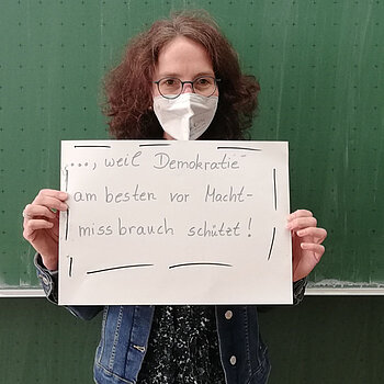 Die Lehrerin Elke Balzhäuser steht vor einer Tafel und hält ein Schild mit der Aufschrift: ..weil Demokratie am besten vor Machtmissbrauch schützt! 
