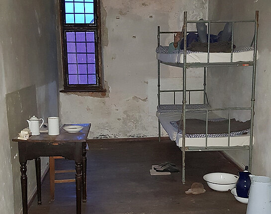 Gefängnsizelle mit schlichtem Stockbett und Nachttopf darunter. An der Wand gegenüber ein kleines Tischchen mit Blechgeschirr.