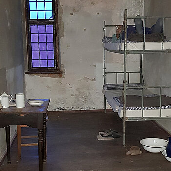 Gefängnsizelle mit schlichtem Stockbett und Nachttopf darunter. An der Wand gegenüber ein kleines Tischchen mit Blechgeschirr.