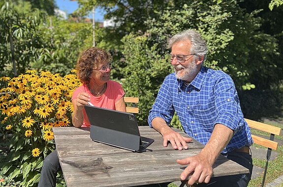 Eine Frau und ein Mann sitzen an einem Gartentisch, im Hintergrund Büsche und gelbe Blumen. Auf dem Tisch ein Laptop 