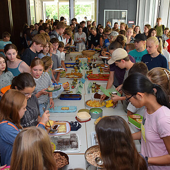 Ein Raum mit vielen Schülerinnen und Schüler, die sich um Tische scharen, auf denen Essen zu sehen ist.