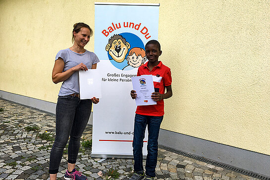 Junge Frau und Junge stehen vor dem Rollup mit dem Balu und Du-Logo und zeigen ihre Urkunden.