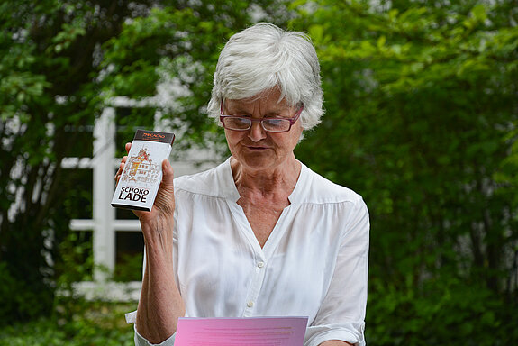 Frau in weißer Bluse mit Brille vorne auf der Nase hält eine Tafel Schokolade hoch.