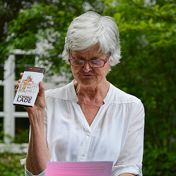 Frau in weißer Bluse mit Brille vorne auf der Nase hält eine Tafel Schokolade hoch.
