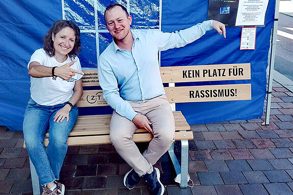 Tabea Janson und Kai Kallbach sitzen auf der Bank "kein Platz für Rassismus". Unter der Schrift gibt es keine Sitzfläche mehr.