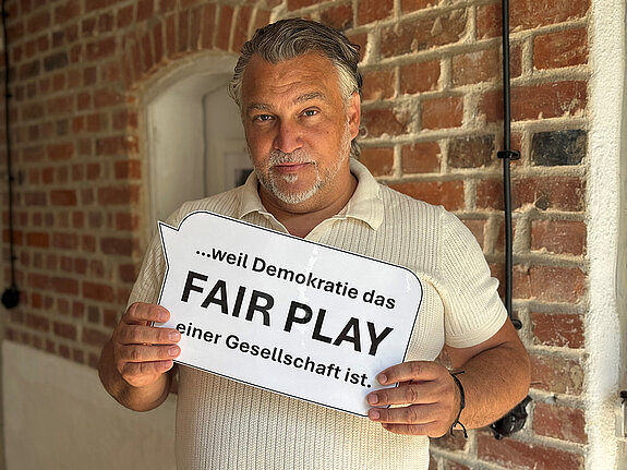 Sebastian Wanzke (Stadtverband Sport) feiert die Demokratie, weil Demokratie das FAIR PLAY einer Gesellschaft ist. Foto: Wanzke Ein Mann im hellen Shirt steht vor einer unverputzten Ziegelwand und hält eine gedruckte Sprechblase in der Hand.