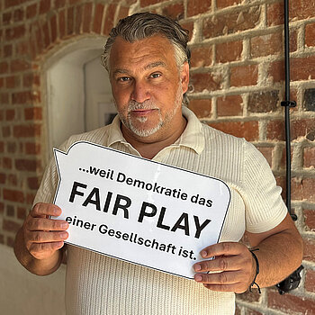 Sebastian Wanzke (Stadtverband Sport) feiert die Demokratie, weil Demokratie das FAIR PLAY einer Gesellschaft ist. Foto: Wanzke Ein Mann im hellen Shirt steht vor einer unverputzten Ziegelwand und hält eine gedruckte Sprechblase in der Hand.