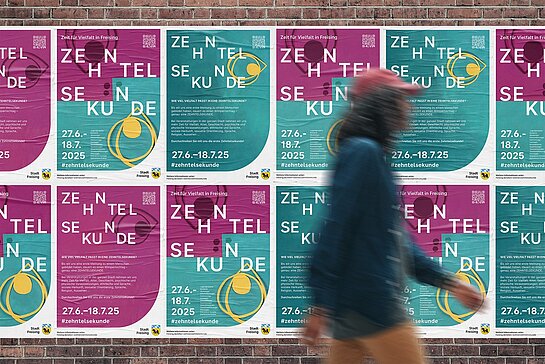 Auf dem Bild ist eine Plakatwand mit einer vorbeilaufenden Person zu sehen.
