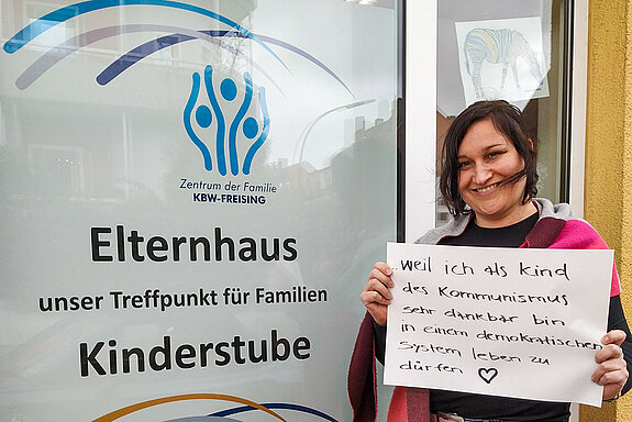 Michaela John steht vor dem Elternhaus und hält ein Schild mit dem Text: "weil ich als Kind des Kommunismus dankbar bin, in einem demokratischen System leben zu dürfen." 