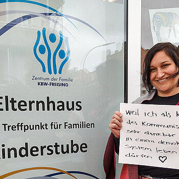 Michaela John steht vor dem Elternhaus und hält ein Schild mit dem Text: "weil ich als Kind des Kommunismus dankbar bin, in einem demokratischen System leben zu dürfen." 