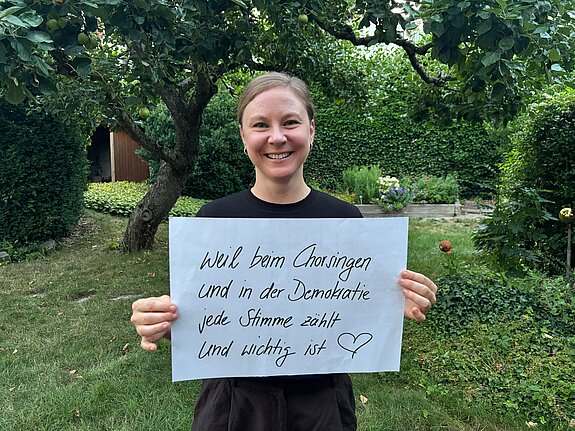Mimi Neumair (Musikerin und Gründerin von Anchora) feiert die Demokratie, weil beim Chorsingen und in der Demokratie jede Stimme wichtig ist. Foto: Mimi Neumair Eine Frau mit dunkelblonden Haaren lacht und steht in einem Garten und hält ein Schild.