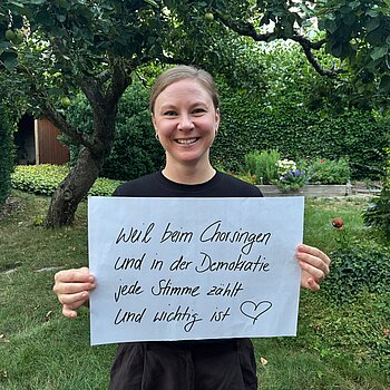 Mimi Neumair (Musikerin und Gründerin von Anchora) feiert die Demokratie, weil beim Chorsingen und in der Demokratie jede Stimme wichtig ist. Foto: Mimi Neumair Eine Frau mit dunkelblonden Haaren lacht und steht in einem Garten und hält ein Schild.