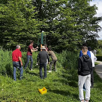 auf einer Grünfläche mit Bäumen im Hintergrund arbeiten zwei Jungen unter Anleitung Erwachsener und rammen einen Pfosten in den Boden.