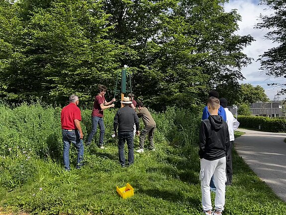 auf einer Grünfläche mit Bäumen im Hintergrund arbeiten zwei Jungen unter Anleitung Erwachsener und rammen einen Pfosten in den Boden.