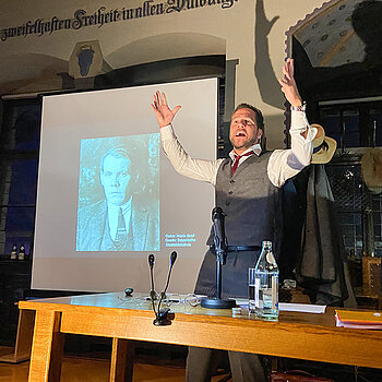 Ein Mann mit erhobenen Händen neben einem auf die Leinwand projizierten Portrait.