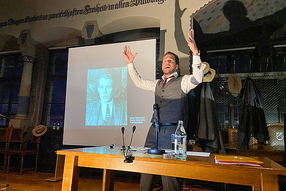 Ein Mann mit erhobenen Händen neben einem auf die Leinwand projizierten Portrait.