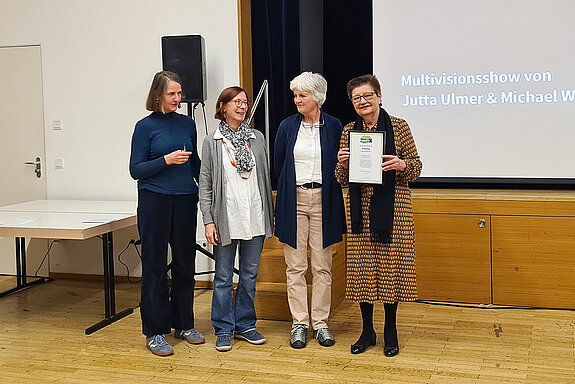4 Frauen stehen vor einer Leinwand. Eine zeigt eine Urkunde