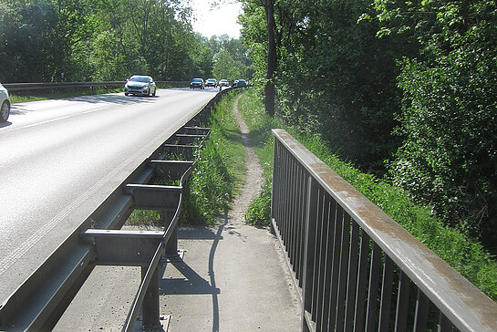 eine Straße, rechts davon das Ende einer Brücke mit einem sehr schmalen Fußweg, der in einen Trampelpfad hinter der Leitplanke mündet