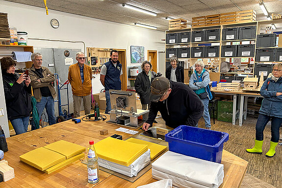 ein Mann arbeitet mit Wachs an einem Arbeistisch. Eine Gruppe steht um ihn herum und beobachtet ihn dabei.