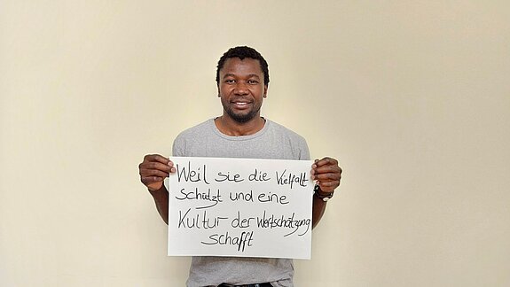 Sem Haikali feiert die Demokratie, weil sie die Vielfalt schützt und eine Kultur der Wertschätzung schafft. Foto: Migrationsrat ein Mann im hellgrauen Shirt steht vor einer hellen Wand und hält ein Schild.
