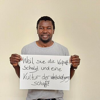 Sem Haikali feiert die Demokratie, weil sie die Vielfalt schützt und eine Kultur der Wertschätzung schafft. Foto: Migrationsrat ein Mann im hellgrauen Shirt steht vor einer hellen Wand und hält ein Schild.