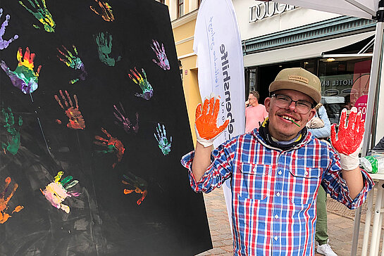Junger Mann mit rot bemalten Händen vor einer schwarzen Tafel mit bunten Handabdrücken.