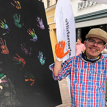 Junger Mann mit rot bemalten Händen vor einer schwarzen Tafel mit bunten Handabdrücken.