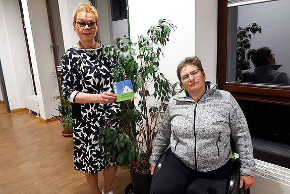 zwei Frauen, davon eine im Rollstuhl vor einer Grünpflanze