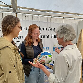 Eine Frau mit kurzen grauen Haaren und einem Ball in der Hand spricht mit zwei jungen Frauen Mitte 20. 