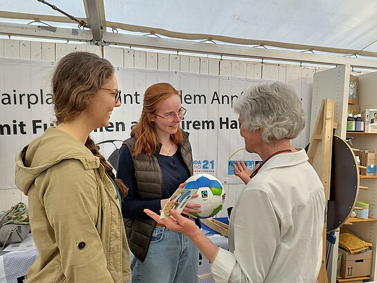 Eine Frau mit kurzen grauen Haaren und einem Ball in der Hand spricht mit zwei jungen Frauen Mitte 20. 