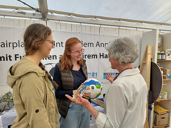 Eine Frau mit kurzen grauen Haaren und einem Ball in der Hand spricht mit zwei jungen Frauen Mitte 20. 