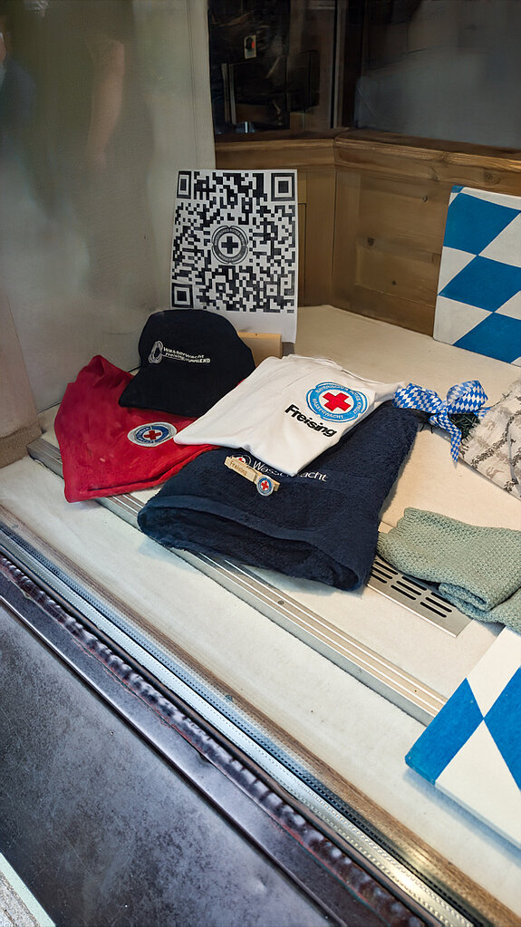 Im Fenster der Dirndltruhe zeigte sich die Wasserwacht. Foto: Stadt Freising in einem Fenster liegen mehrere Wasserwacht-Shirts sowie weiß-blaue Rauten und ein QR-Code.