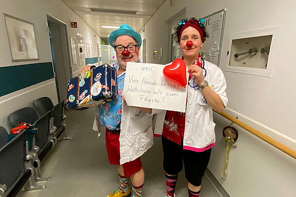 Die Klinkclowns Paletti und Schmatz feiern die Demokratie, weil vier Hände besser klatschen als zwei Fäuste. Foto: Klinikclowns Ein Mann und eine Frau als Clowns maskiert und mit weißen Kitteln stehen in einem Klinik-Gang.