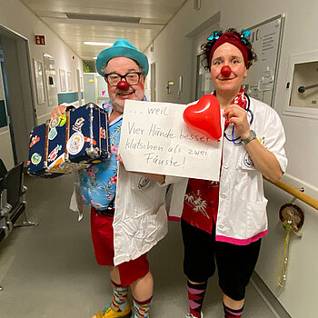 Die Klinkclowns Paletti und Schmatz feiern die Demokratie, weil vier Hände besser klatschen als zwei Fäuste. Foto: Klinikclowns Ein Mann und eine Frau als Clowns maskiert und mit weißen Kitteln stehen in einem Klinik-Gang.