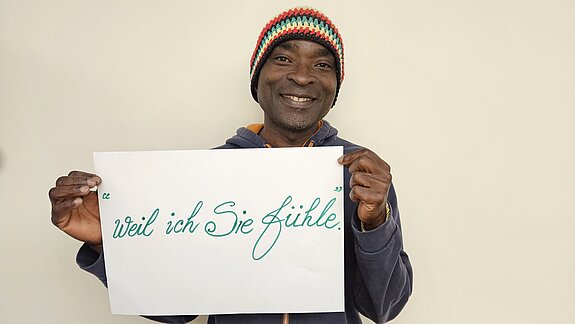 Pepito Anumu feiert die Demokratie, weil er sie fühlt. Foto: privat Ein Mann mit bunter Strickmütze hält lachend ein Schild. Die Schrift ist groß und schön geschwungen.