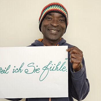 Pepito Anumu feiert die Demokratie, weil er sie fühlt. Foto: privat Ein Mann mit bunter Strickmütze hält lachend ein Schild. Die Schrift ist groß und schön geschwungen.