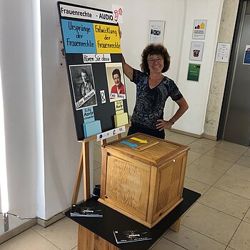 Wahlurne mit Plakat und einer Frau, die das präsentiert.
