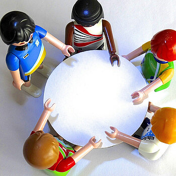 sind fünf Playmobil Figuren von oben zu sehen, die um einen runden Tisch stehen. Die Figuren haben unterschiedliche Haut- und Haarfarben.