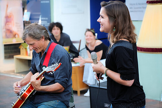 Ein Mann mit Gitarre sitzend, eine Frau stehend sehen nach links. Im Hintergrund Publikum.