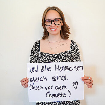 Stadtratsmitglied Lucy Knott. Foto: Stadt Freising Eine Frau in einem schwarzen Kleid mit weiß/gelben Blumen zeigt ein Schild mit der Aufschrift "Weil alle Menschen gleich sind (Auch vor dem Gesetz)