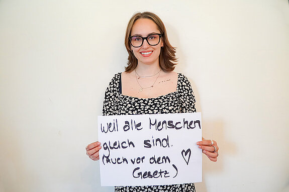 Stadtratsmitglied Lucy Knott. Foto: Stadt Freising Eine Frau in einem schwarzen Kleid mit weiß/gelben Blumen zeigt ein Schild mit der Aufschrift "Weil alle Menschen gleich sind (Auch vor dem Gesetz)
