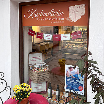 Schaufenster mit einem roten Tisch davor, Blumenstock darauf. Im Fenster ein Stapel Pizzakartons. Der oberste geöffnet mit eine Pizza darin, Playmobil-Männchen und Fähnchen