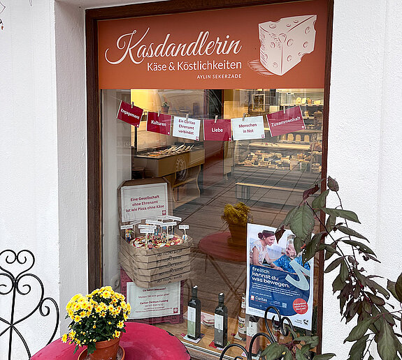 Schaufenster mit einem roten Tisch davor, Blumenstock darauf. Im Fenster ein Stapel Pizzakartons. Der oberste geöffnet mit eine Pizza darin, Playmobil-Männchen und Fähnchen