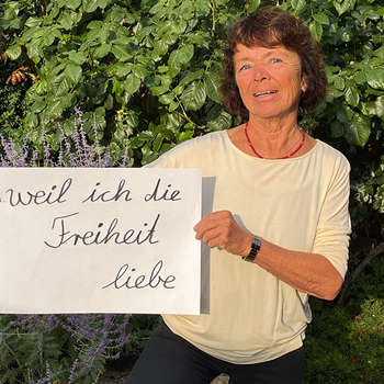 Marita Hanold (Agenda21 und Treffpunkt Ehrenamt) feiert die Demokratie, weil sie die Freiheit liebt. Foto: Marita Hanold Eine Frau vor einer grünen Hecke im hellen Oberteil und mit einem Schild.