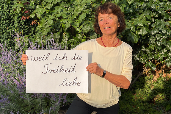 Marita Hanold (Agenda21 und Treffpunkt Ehrenamt) feiert die Demokratie, weil sie die Freiheit liebt. Foto: Marita Hanold Eine Frau vor einer grünen Hecke im hellen Oberteil und mit einem Schild.