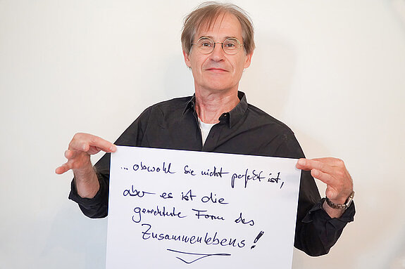 Stadtratsmitglied Manfrd Drobny. Foto: Stadt Freising Ein Mann in einem schwarzen Hemd zeigt ein Schild mit der Aufschrift "obwohl sie nicht perfekt ist,aber es ist die gerechteste Form des Zusammenlebens!"