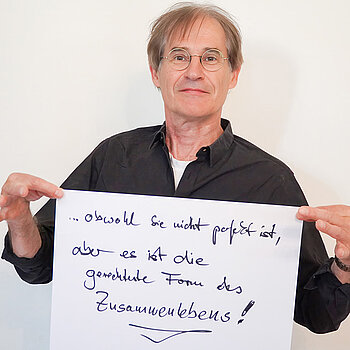 Stadtratsmitglied Manfrd Drobny. Foto: Stadt Freising Ein Mann in einem schwarzen Hemd zeigt ein Schild mit der Aufschrift "obwohl sie nicht perfekt ist,aber es ist die gerechteste Form des Zusammenlebens!"