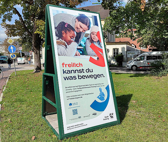 freilich kannst Du was bewegen.. die Engagement-Datenbank. Foto: Stadt Freising ein übermannsgroßer Aufsteller im Grünen mit einer Frau, die sich lachend zu einem Kind beugt.