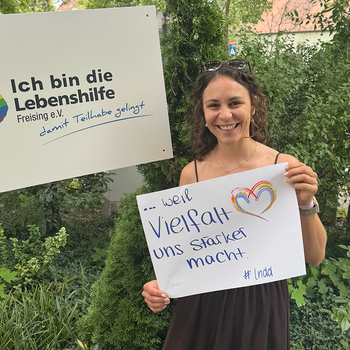 Alexandra Deml feiert die Demokratie weil Vielfalt uns stärker macht. Foto: Lebenshilfe Eine junge Frau mit Brille im Haar steht im Grünen und hält ein Schild, daneben hält jemand ein Schild "Ich bin die Lebenshilfe Freising e.V."